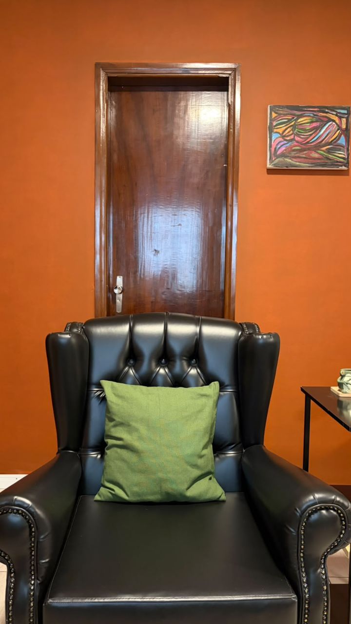Consultório da Jade Teixeira: poltrona de couro preta com almofada verde diante de uma parede terracota, com porta de madeira e um quadro colorido