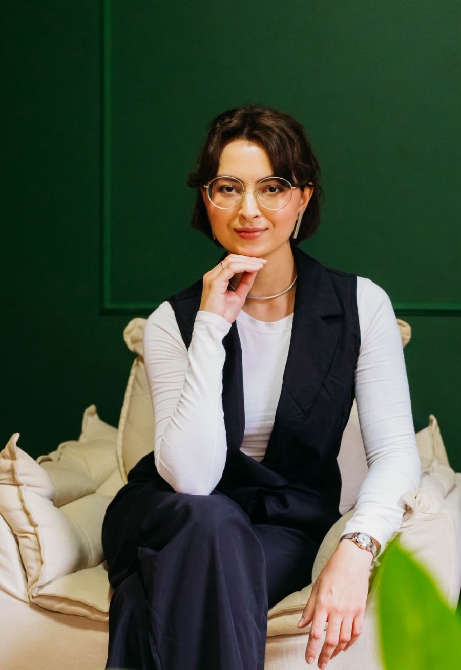 Jade Teixeira sentada em um sofá claro diante de uma parede verde, com a mão no queixo e olhar tranquilo